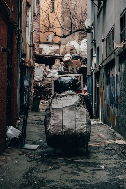 A narrow urban alleyway scene featuring a large, wheeled rubbish bag made of weathered, beige fabric positioned centrally in the foreground, with visible creases and dirt markings. Behind it, a pile of mixed waste cardboard boxes, plastic bags, and other discarded materials is stacked unevenly against a wooden pallet and a backdrop of older brick and concrete buildings. The alley’s ground is paved with dark asphalt, showing signs of dirt and debris, and the surrounding walls are covered in graffiti and peeling paint, indicating a neglected environment. To the right, a chain-link fence with some metal bars and electrical wiring run along the building's exterior, while on the left, a rusted metal door and a small trash bin are present. The lighting appears natural, possibly overcast, casting soft shadows across the scene, and the overall atmosphere suggests a location where private waste collection services, such as those provided by Rubbish Removal Tooting, might be needed for clearing accumulated rubbish in an alleyway often used for informal waste disposal or storage.