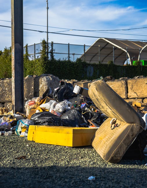 A narrow urban alleyway scene featuring a large, wheeled rubbish bag made of weathered, beige fabric positioned centrally in the foreground, with visible creases and dirt markings. Behind it, a pile of mixed waste cardboard boxes, plastic bags, and other discarded materials is stacked unevenly against a wooden pallet and a backdrop of older brick and concrete buildings. The alley’s ground is paved with dark asphalt, showing signs of dirt and debris, and the surrounding walls are covered in graffiti and peeling paint, indicating a neglected environment. To the right, a chain-link fence with some metal bars and electrical wiring run along the building's exterior, while on the left, a rusted metal door and a small trash bin are present. The lighting appears natural, possibly overcast, casting soft shadows across the scene, and the overall atmosphere suggests a location where private waste collection services, such as those provided by Rubbish Removal Tooting, might be needed for clearing accumulated rubbish in an alleyway often used for informal waste disposal or storage.