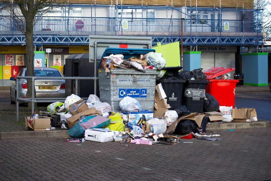 In the foreground, a large collection of mixed waste, including black and red wheelie bins, cardboard boxes, paper bags, and miscellaneous refuse, is piled on a paved area adjacent to a curb. The black bin with white lettering and the red bin are overflowing with refuse, with some items spilling onto the ground. The pile comprises crumpled, flattened, and partially opened cardboard boxes, plastic bags filled with rubbish, and loose papers and packaging scattered across the pavement. Behind the waste collection, there are parked cars, with one silver vehicle partially visible on the left side, featuring a yellow registration plate. Further back, a building with a yellow storefront and glass windows has signs indicating commercial premises, with a blue scaffold structure covering the upper level. The environment appears urban, and the scene suggests an instance of irregular rubbish disposal, possibly handled through private waste collection services like those offered by Rubbish Removal Tooting, to support alternative waste handling outside of standard municipal rubbish collection. The scene is captured during daylight, under natural lighting, with no people visible in the image.
