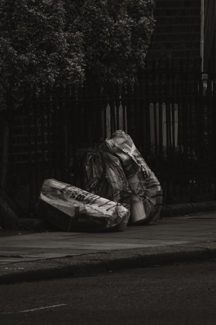 The image depicts a narrow urban alleyway cluttered with a large, weathered black and gray wheeled rubbish bin in the foreground, positioned centrally on the damp, uneven asphalt surface. Behind the bin, there is an extensive pile of discarded cardboard boxes, plastic bags, and other waste materials stacked haphazardly against a backdrop of aged, multi-storey buildings with brick and plaster facades. The alley is bordered by dark, graffiti-covered walls on the right, while the left side features a rusty metal gate and a weathered door. Overhead, leafless tree branches extend across the scene, indicating a winter setting, and the overall atmosphere is dimly lit with muted natural light filtering through. This setting illustrates typical private waste disposal or on-site clearance within an urban environment, aligning with independent rubbish removal services such as those provided by Rubbish Removal Tooting, showcasing the type of waste handled outside of conventional council collections.