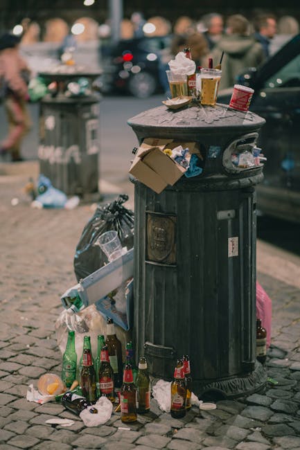 A black, cylindrical rubbish bin with a domed lid, situated on a cobblestone pavement in an urban area. The top of the bin holds discarded food and drink items, including paper cups with plastic lids and straws, a crumpled takeaway box, and a plastic cup. Attached to the side of the bin is a partially open cardboard container, with its contents spilling over. Surrounding the base of the bin, there are numerous discarded glass bottles, some broken, along with crumpled paper and other litter. Behind the bin, a green public trash or recycling bin is visible in the background, along with blurred figures of pedestrians and parked vehicles, suggesting a busy city street setting. A grey car is partially visible to the right of the trash bin, adding to the urban environment context. The scene is lit with natural daylight, emphasizing the clutter and informal disposal of refuse common in busy public areas, which relates to independent rubbish collection services nearby.