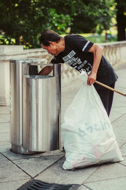 A man with short dark hair, wearing a dark T-shirt with white printed text, is seen disposing of rubbish into a metallic public litter bin on a paved sidewalk. The bin is cylindrical, made of stainless steel with a smooth, shiny finish, and has two compartments with openings at the top. The man is slightly bent forward, holding a white plastic bag filled with waste in his left hand while using a wooden stick or grabber in his right hand to assist in placing the rubbish into the bin. The background features a stone balustrade and lush green foliage, indicating an outdoor urban park or street setting. The scene is well-lit with natural daylight, highlighting the metallic surface of the litter bin and the overall cleanliness of the area, illustrating a typical scene of independent rubbish disposal supported by waste management services such as Rubbish Removal Tooting.