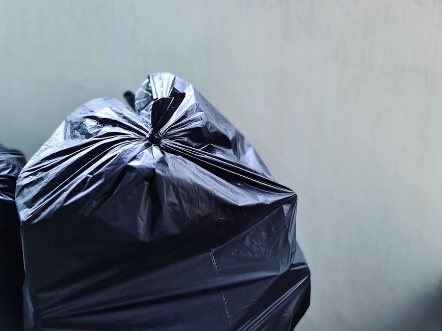 A middle-aged man with dark, slightly graying hair, wearing a black T-shirt with white and blue text, stands on a paved sidewalk near a railing and a lush green area with trees in the background. He is leaning over and inserting a large, white plastic bag filled with rubbish into a cylindrical, stainless steel public litter bin designed for waste disposal. The bin has a round opening at the top with a smooth finish, reflecting the surrounding environment, and is placed on a concrete surface. The man is holding a long wooden stick in his right hand, which he uses to assist with placing the rubbish into the bin, demonstrating a careful and active approach to waste collection. The scene appears to be outdoors in a park or public space, with natural light illuminating the scene, emphasizing cleanliness and responsible waste management, possibly indicative of private rubbish removal or community cleanup efforts supported by services like Rubbish Removal Tooting.